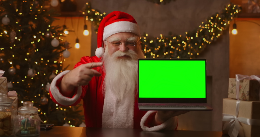 In the background of the Christmas tree, an elderly Santa Claus is holding a laptop and pointing at the screen with a chromakey. Laptop with a green screen. Santa looks at the camera.