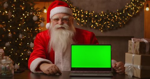 In the background of the Christmas tree, an elderly Santa Claus is holding a laptop and pointing at the screen with a chromakey. Laptop with a green screen. Santa looks at the camera.