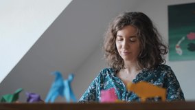 A young woman having fun folding paper to create Origami figures in living room. Enjoying the peaceful and relaxing traditional Japanese art. Handmade colorful animals on wooden table. Slow-motion 4K. - Powered by Shutterstock - Get 15% off with code: PIKWIZARD15