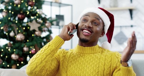 African American happy man sit near decorated glowing tree wearing santa hat enjoying distant talk with friend on Christmas Eve on smartphone, receive congratulations best wishes on New Year concept