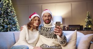 Joyful family couple talking on video chat online on smartphone and waving hands while sitting at home with glowing xmas tree Caucasian husband and wife greeting friends on video call New Year concept