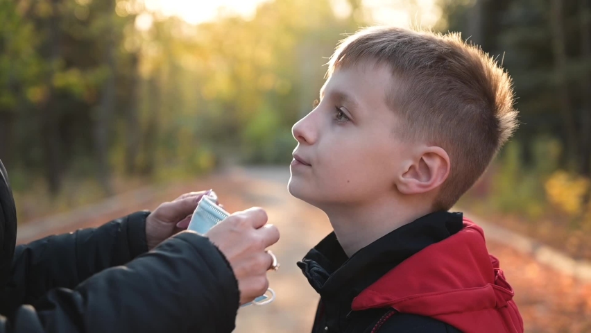Close up of young woman put medical mask on face of little cute boy and hugs him. Protect from virus, take care of health of child outdoors, modern family life in pandemic epidemic of respiratory