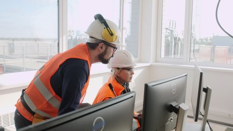 Skilled employees in orange uniform and helmets work on computer in hall at contemporary electrical distribution station office