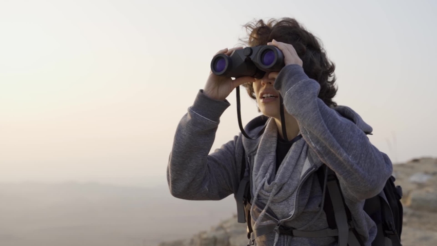 Excited female traveler or birdwatcher smiles and enjoys looking with binoculars. Backpacker or hiker standing on top of desert mountain or hill watching the epic view during sunset or sunrise