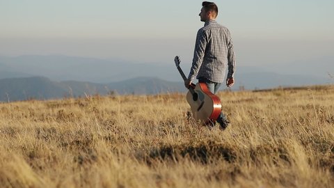 Man with acoustic instrument enjoying nature and sunlight. People, lifestyle, student, expression and style concept. Man in a shirt with guitar going along the field on the beautiful sunset.