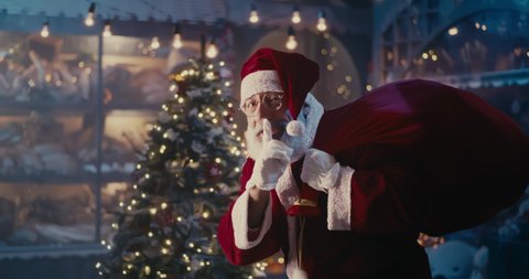 Cheerful Santa Claus with bag of presents smiling and asking to keep silence while walking near decorated Christmas tree at night
