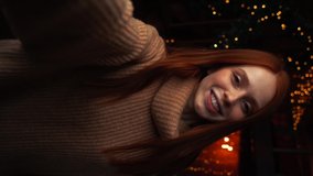 Close-up POV of cheerful young woman greeting and congratulating relatives with Merry Christmas by video call, background of xmas tree at cozy living room with festive interior, vertical orientation.