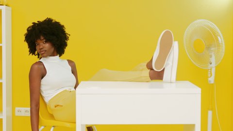 Portrait of stylish African woman with black curly hair, looking at camera, sitting at desk in yellow room, holding legs on table, fan on pedestal is on, creative space, Slow motion.