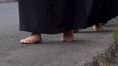 People in black robes walking barefoot on an asphalt street accompanied by a man in black boots