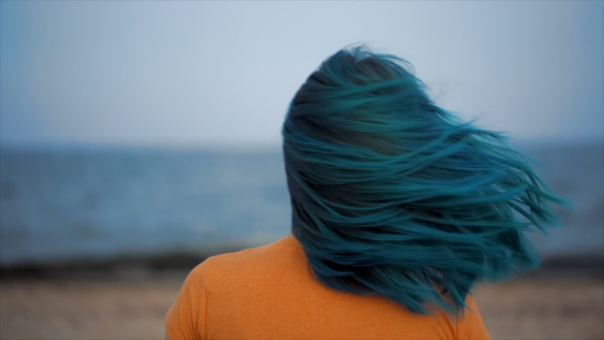 Unusual woman shakes her blue dyed hair on sea beach background. Portrait of hipster girl with unique fashionable hairstyle, she enjoying summertime alone, demonstrate her fresh color. Royalty-Free Stock Footage #1063058497