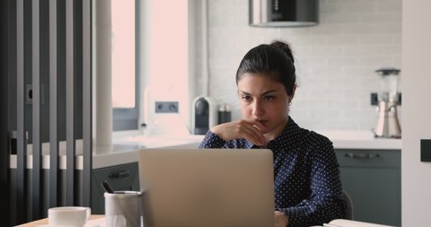 Focused young mixed race indian woman sitting at desk in modern studio apartment, working remotely on computer, pondering thinking of problem solution, preparing electronic report alone indoors.