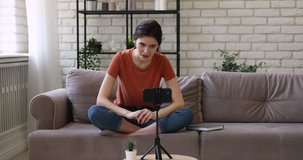 Beautiful millennial female blogger sitting on comfortable couch in front of mobile phone on tripod, recording talking video for personal channel, sharing life news or filming vlog alone at home.