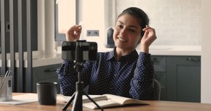 Smiling millennial indian ethnic woman sitting in front of mobile phone on tripod, recording educational video or making college online project task, sharing knowledge, streaming webinar for followers