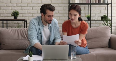 Focused young married couple sitting on sofa with paper documents, discussing banking mortgage money loan agreement, apartment purchase contract terms of conditions, family financial affairs.