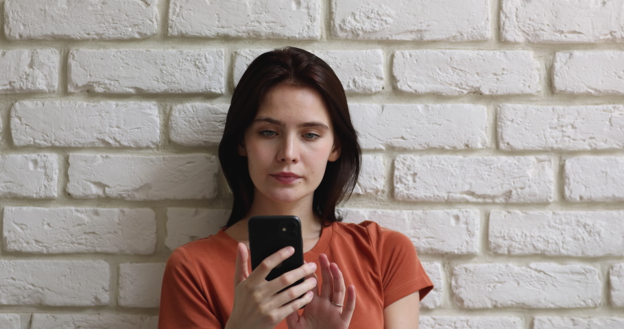 Head shot serious young pretty caucasian girl standing near brick wall, using mobile phone applications, web surfing information, involved in online shopping or communicating distantly with friends. Royalty-Free Stock Footage #1063059136