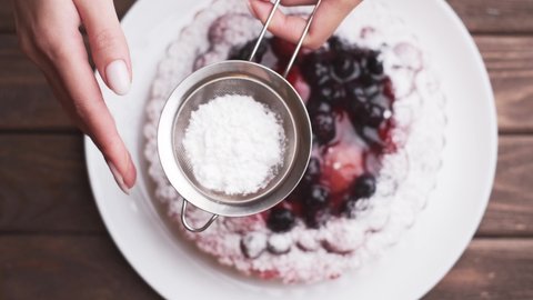 Female hands of chef with metal sieve sprinkling a cake with powdered sugar at pastry shop kitchen, top view.