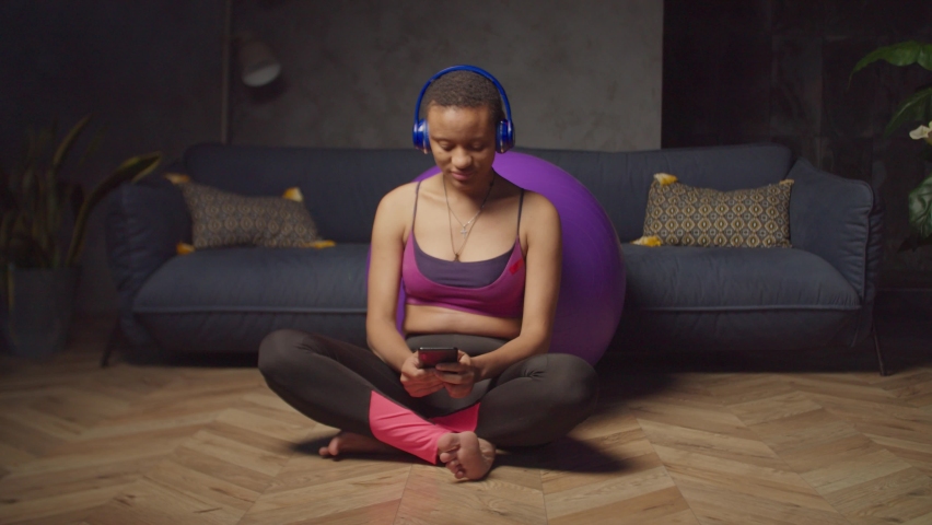 Positive charming sporty fitness african woman in wireless headphones sitting cross legged on floor, text messaging on smartphone while relaxing and chatting online after indoor workout.
