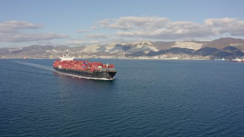 Aerial footage of ultra large container ship at sea, side view. Cargo ship with colourful containers leaves port in sunny weather. Following drone 4k footage sea port at background