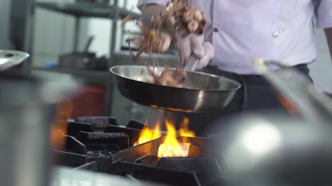Skilled chef in uniform cooks tasty fresh octopuses in large frying pan on stove with burning flame in luxury seafood restaurant kitchen extreme close view.