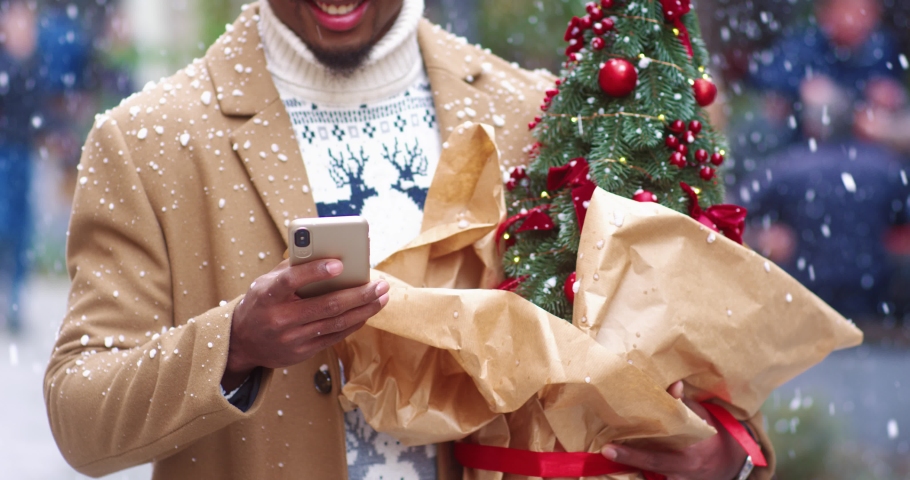 Handsome African American man in stylish coat is typing message on smartphone holding small Christmas tree standing on street under snowfall.