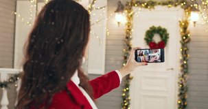 Back view of young female videochatting on cellphone on winter holiday with female mixed-races friends and grey-haired dad while standing in snowy decorated city on New Year. Christmas mood. Close up