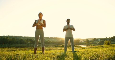 Male and female cacasian yogis standing on green grass early in morning and rising hands up. Man and woman doing yoga and sun salutation asana in nature. Couple breathing and relaxing outdoor.