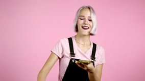 Pretty woman with happy face scatters, waste money. Hipster girl with violet dyed hair overspend US currency. Lady is flush with dollars on pink studio background .