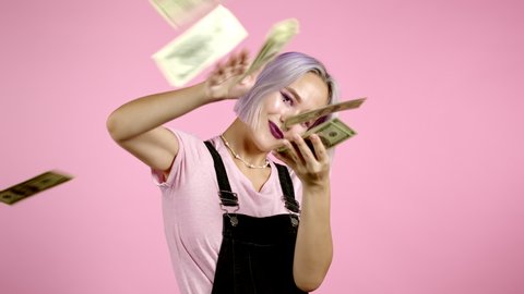 Pretty woman with happy face scatters, waste money. Hipster girl with violet dyed hair overspend US currency. Lady is flush with dollars on pink studio background .