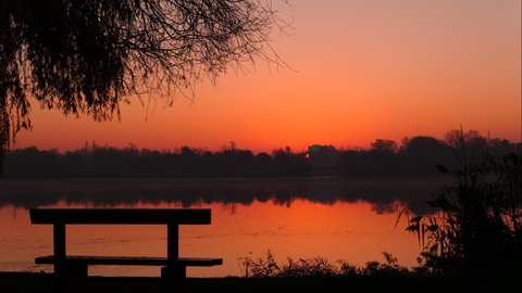 Amazing sunrise over the water. Time lapse with close up of the sun on the horizon. Lake and silhouette of weeping willow with isolated bench in the foreground.