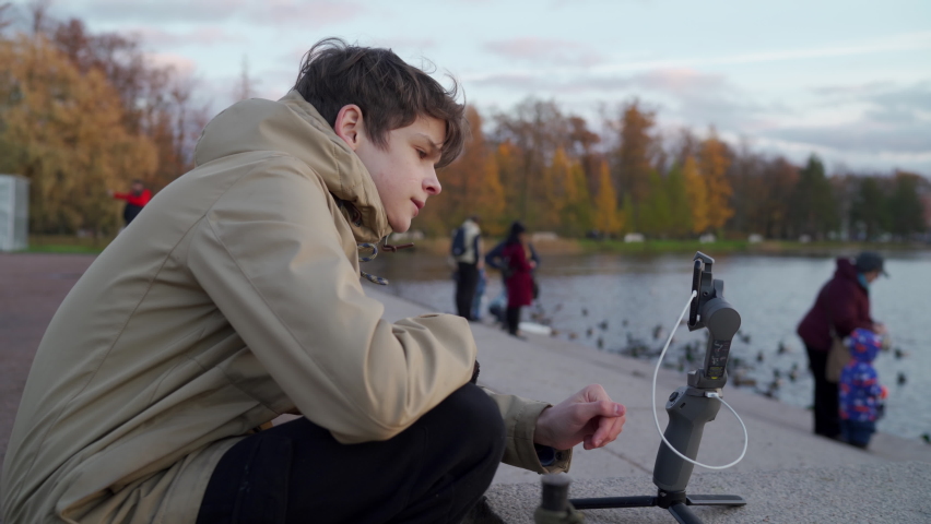 Vlogger boy with mobile phone camera stabilizer recording outdoor video blog in autumn park. Teenager shooting video for travel vlogging. Catherine Park, Tsarskoye Selo, Saint Petersburg, Russia.