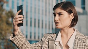 Attractive female lawyer with coffee to go making video call with customers on smartphone outdoor. Young businesswoman in downtown