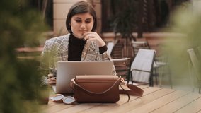 Young positive female lawyer talking on video call with customers and making notes in diary during coffee break outdoor. Businesswoman with laptop