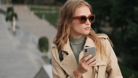 Beautiful stylish student girl wearing sunglasses and trench coat taking selfie on smartphone on street