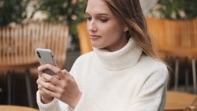 Beautiful student girl in white cozy sweater texting with friends in online chat on smartphone on street