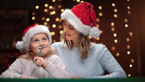 Mother and daughter in Santa Claus festive hats writing wish list Christmas presents together. Happy female child and parent enjoying Xmas eve night at holiday atmosphere. Medium shot on RED camera
