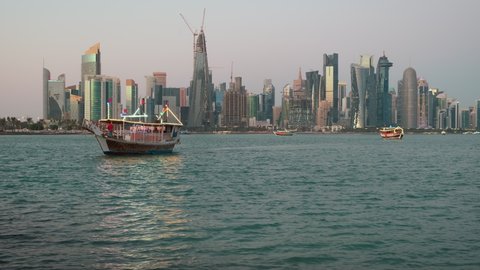 Doha skyline from the corniche promenade sunset zooming in shot showing dhow with Qatar flags in the Arabic gulf  in foreground and clouds in the sky in background