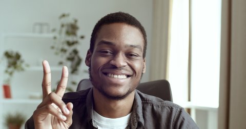 Portrait of smiling black male african guy businessman student sitting at table in office at home looking at camera showing two fingers second number peace gesture approval friendliness greeting hello