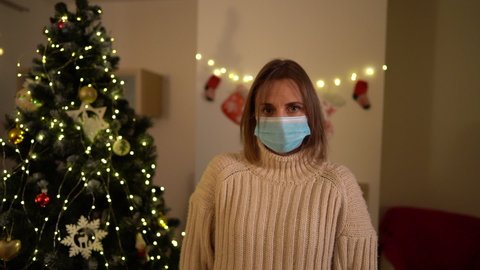 Christmas and covid-19, social distance. Portrait of lonely woman wearing medical mask with sad emotion against the background of a Christmas tree. Coronavirus pandemic