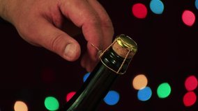 Male hand open a bottle of Christmas champagne close-up. First, unscrew the wire, then remove the plug. A festive night and a solemn moment came. New Year colored lights on a black background. 4K.