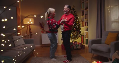 Portrait of cheerful smiling Caucasian married senior couple in decorated room celebrating New Year together in evening. Wife and husband dancing near glowing xmas tree on Christmas. Holidays concept
