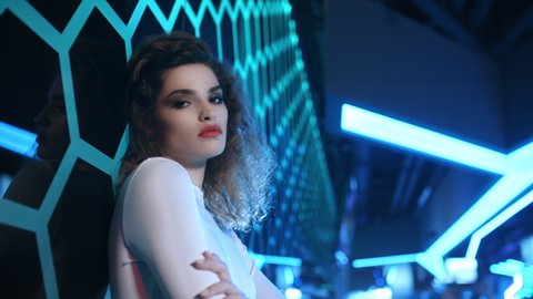 Portrait of beautiful trendy woman wear white outfit posing and looking into camera standing by illuminated wall in night club. Futuristic style, sensual look clubber in neon blue light