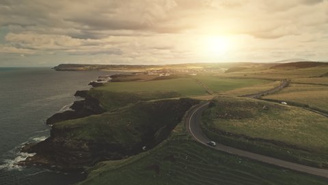 Sun green fields, meadows aerial. Cars drive at road. Ireland hills, farmlands at cloudy day. Nobody nature landscape of Antrim County, United Kingdom, Europe. Cinematic footage drone shot
