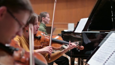 Focus on a young woman playing the violin with a string quartet and grand piano. Selective focus