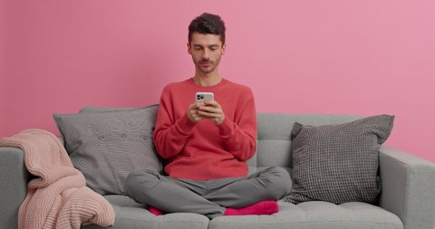 Happy smiling European man sits in lotus pose on comfortable sofa uses mobile phone for online communication surfs social networks poses in modern apartment connected to high speed internet.