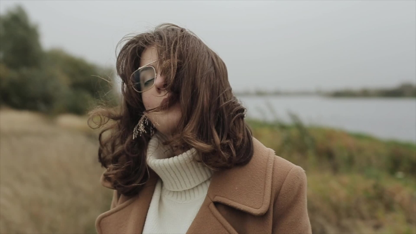 Sensual young woman in glasses stands with closed eyes on the river bank on a windy autumn day and holds a blade of grass in her mouth. Slow motion