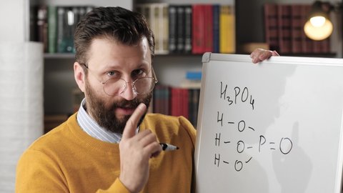 Online education and remote learning concept. Male chemistry teacher talking and looking at camera and writing phosphoric acid formula on white magnetic whiteboard while explaining to camera