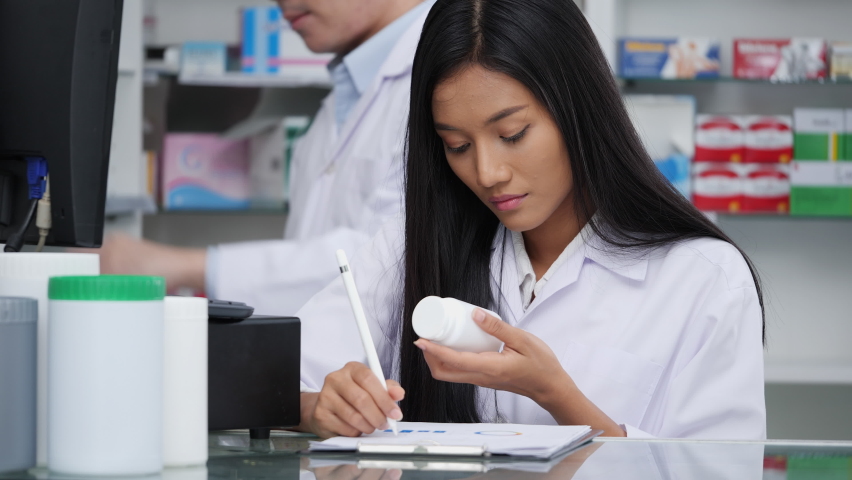 Asian pharmacists working and checking drug in pharmacy store at hospital. Asian doctor checking medicine cabinet and wearing white medical coat. Shelves pharmacy background in drugstore. | Shutterstock HD Video #1063104922