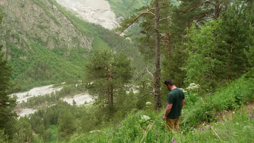 Man stands on green grass and plants in mountains and happily spreads his hands. Happy people