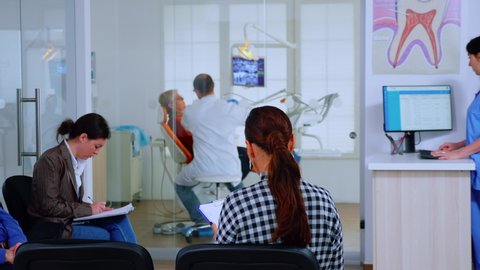 Back view of woman filling in dental document sitting on chiar in waiting room preparing for teeth exemination while doctor working in background. Concept of crowded professional orthodontist office.