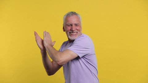 Cheerful funny elderly gray-haired mustache bearded man in casual violet t-shirt isolated on yellow color background studio. People lifestyle concept. Dancing clenching fists waving clapping hands